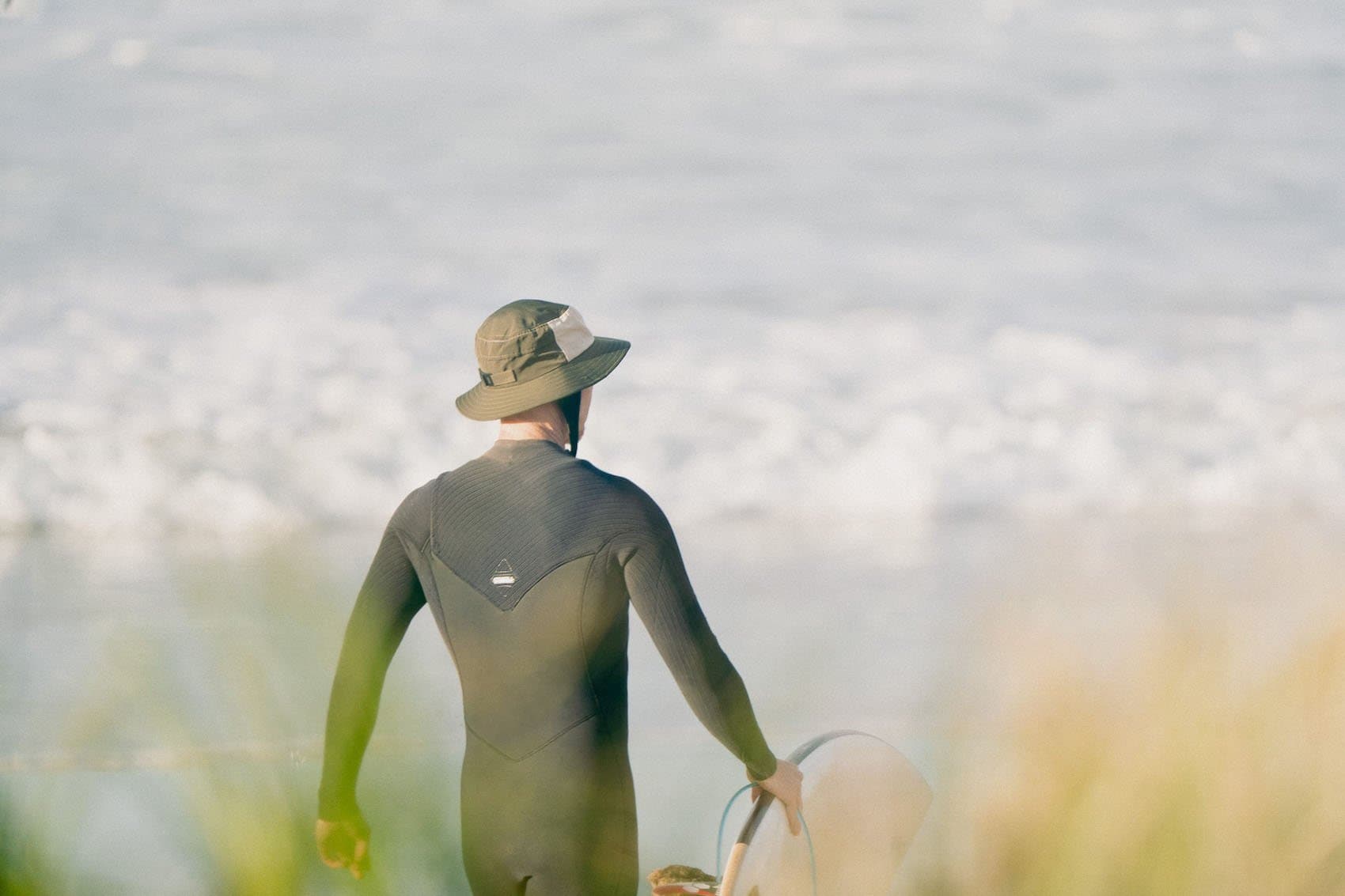 A surfer in an olive OCEANlab hat walking down to the beach, board under arm