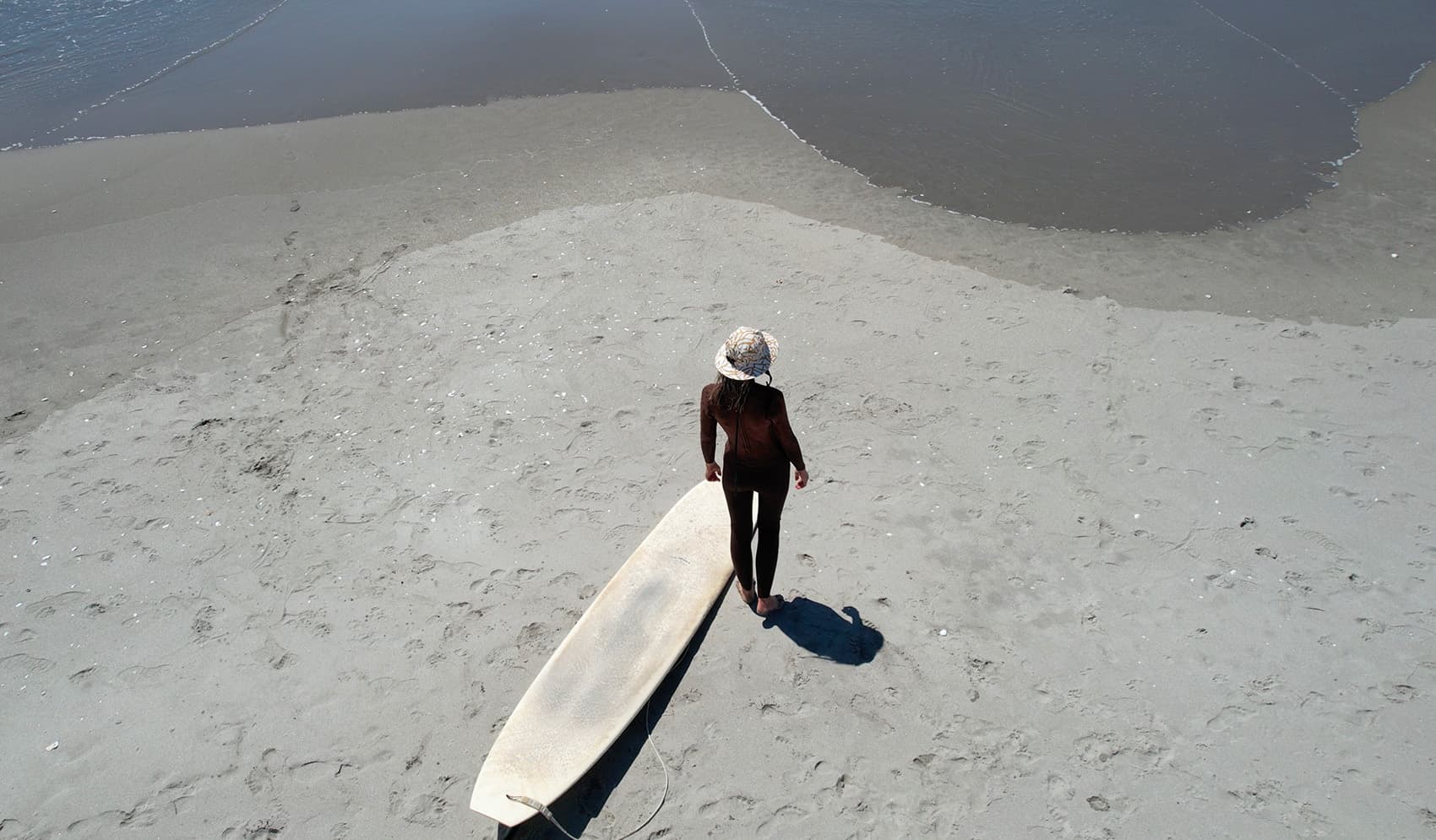 Aerial view of a surfer in an OCEANlab hat standing on the sand with a longboard
