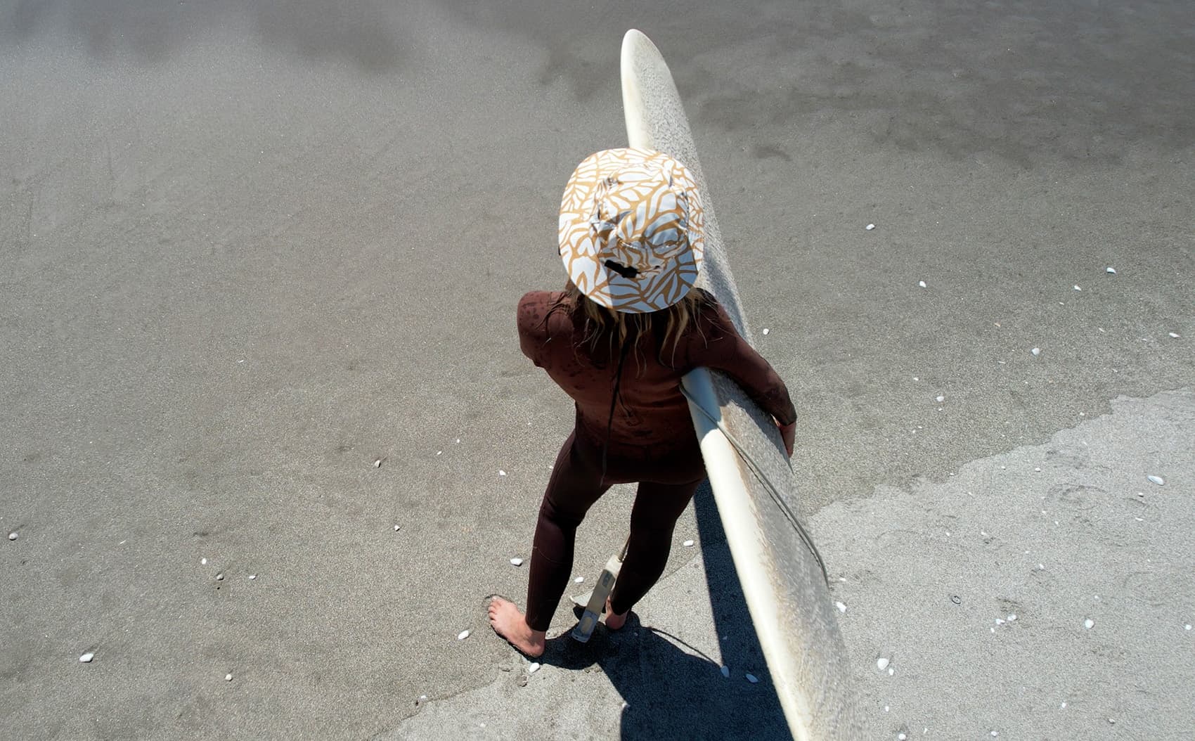 Overhead view of a surfer in the Golden Flow OCEANlab hat carrying a longboard on the sand