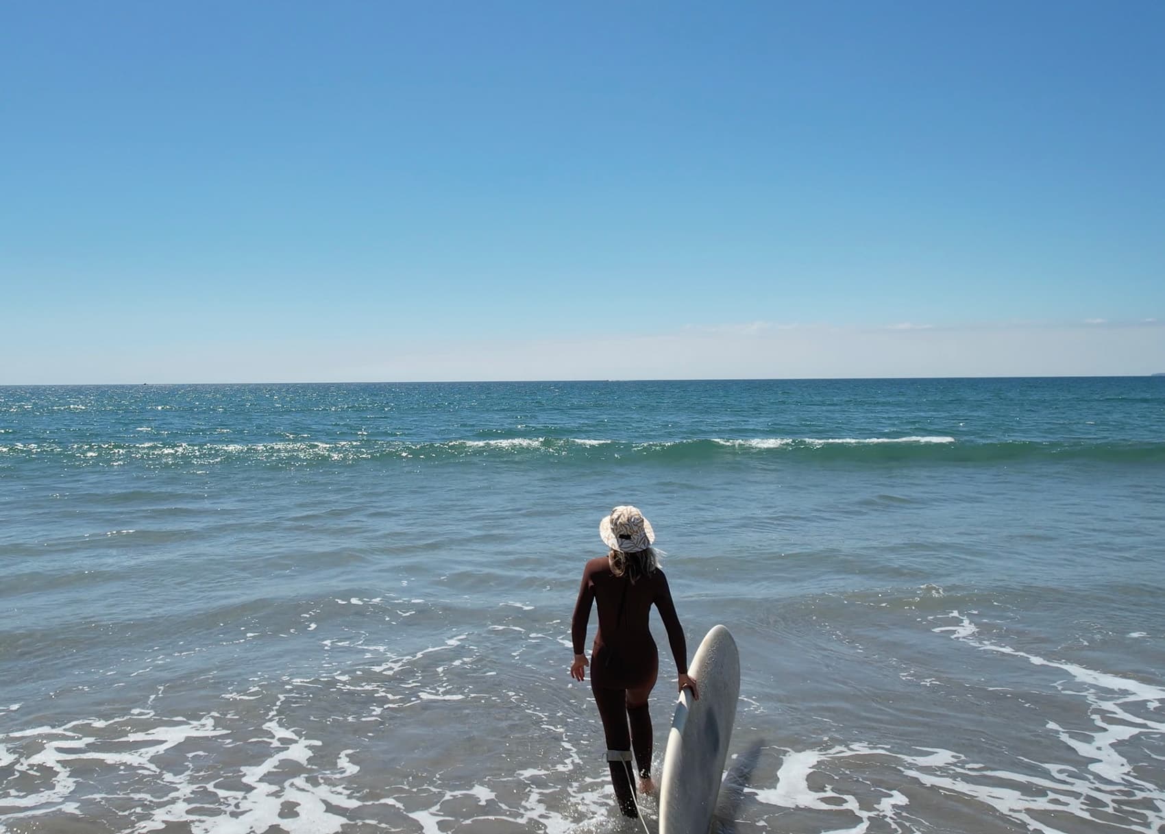 Surfer in an OCEANlab hat and wetsuit walking into the water with a longboard