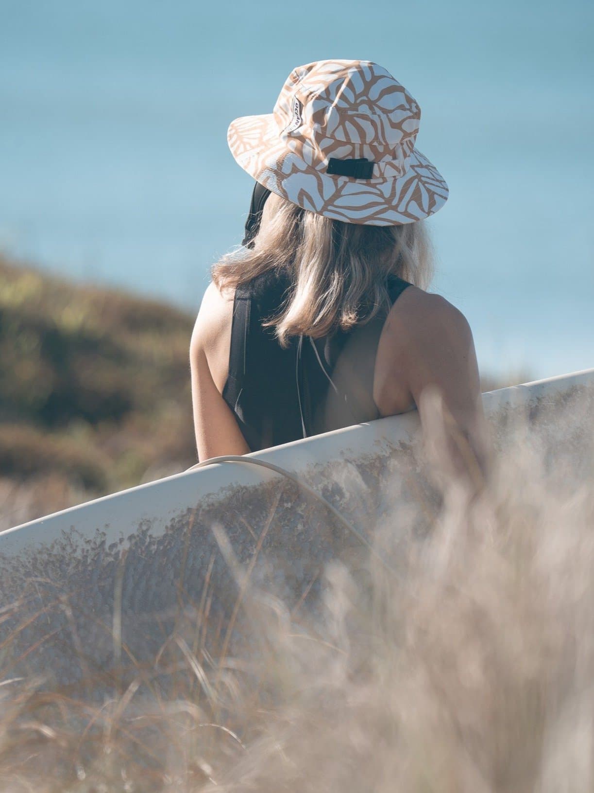 Surfer wearing the Golden Flow surf hat, board under arm, looking out to sea